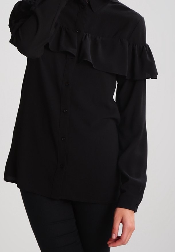 Personne portant un chemisier noir à manches longues boutonné avec un détail à volants sur la poitrine, associé à un pantalon noir.