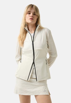 Cream-colored zip-up jacket with a high collar, featuring a textured geometric pattern and black zipper accents. Worn over a white top and skirt.