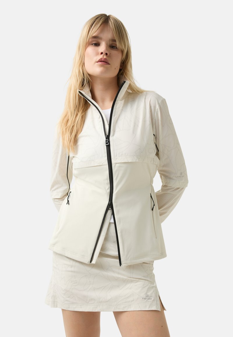 Cremfarbener Zip-up-Jacke mit hohem Kragen, ausgestattet mit einem strukturierten geometrischen Muster und schwarzen Reißverschlussakzenten. Getragen über einem weißen Oberteil und einem Rock.