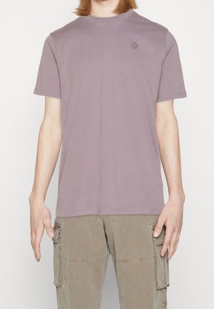 Person iført en ensfarvet mauve kortærmet t-shirt og lysebrune cargo-bukser, stående med armene afslappet ned langs siden.