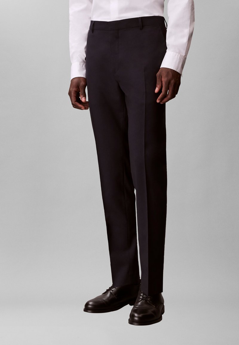 Vyras dėvi juodus, pasiūtus pagal užsakymą, kostiuminės kelnės, juodus odinius batelius su raišteliais ir baltą ilgomis rankovėmis marškinius, stovėdamas priešais paprastą pilką foną.