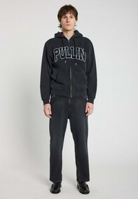 Haut à zip noir avec "PULLIN" en lettres blanches, associé à un jean sombre et des chaussures noires. Dispose de poches avant et d'une capuche à cordon.