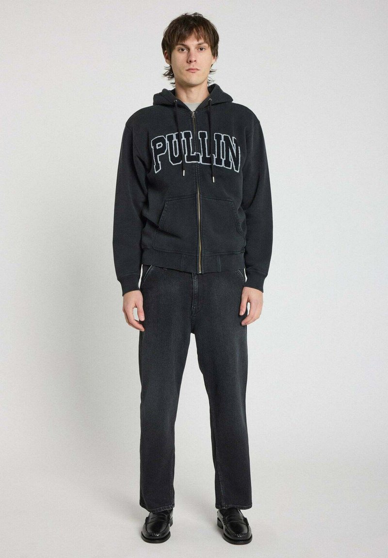 Haut à zip noir avec "PULLIN" en lettres blanches, associé à un jean sombre et des chaussures noires. Dispose de poches avant et d'une capuche à cordon.