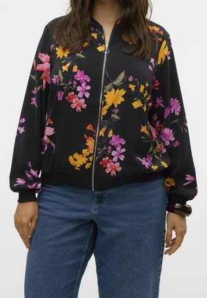 Femme portant une veste noire zippée avec des motifs floraux roses, jaunes et orange et un jean bleu, avec un bracelet sombre au poignet.