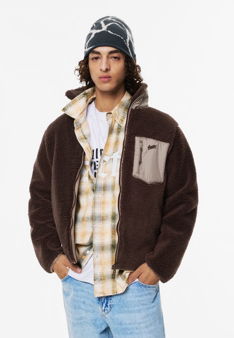 Brązowa polarowa kurtka z zamkiem błyskawicznym w beżowej kieszeni na piersi, nałożona na wykroczoną koszulę w kratkę i białą koszulkę, w połączeniu z jasnoniebieskimi dżinsami.