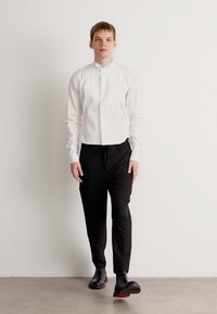 Balta garām piedurknēm krekls ar stāvošu apkakli, apvienots ar melnām pieguļošām biksēm un elegantiem melniem apaviem ar sarkaniem zole.