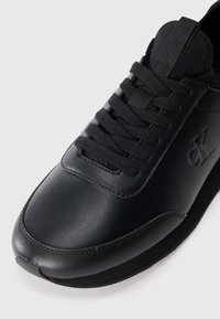 Zwart leren sneaker met een gladde afwerking, voorzien van veters, een gewatteerde tong en een logo aan de zijkant. De zool is vlak en textuur.