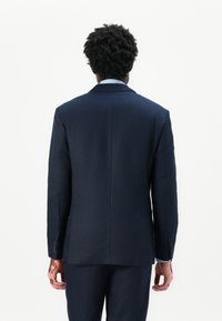 Person mit lockigen dunklen Haaren und Vitiligo, die einen marineblauen Anzug und ein hellblaues Hemd trägt, steht mit dem Rücken zu einem schlichten weißen Hintergrund.
