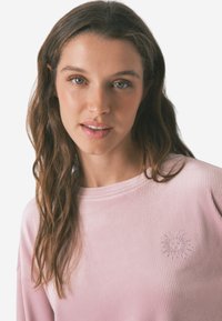 Lichtroze geribbelde long sleeve shirt met een reliëf zon ontwerp op de linkerborst. Soepele textuur en een ontspannen pasvorm.