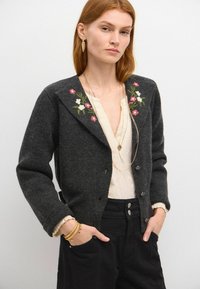 Kardigan in Anthrazitgrau mit floralem Stickerei an den Schultern, durchgehender Knopfleiste und weicher Textur, kombiniert mit einer hellen Bluse und schwarzen Hosen.