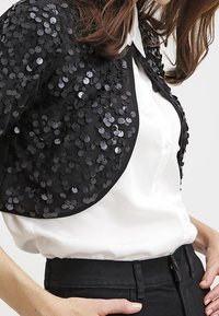 Černý plášť z flitrů s lesklými kulatými flitry, nošený přes bílou košili s límečkem. Má zaoblený tvar a hladkou texturu.