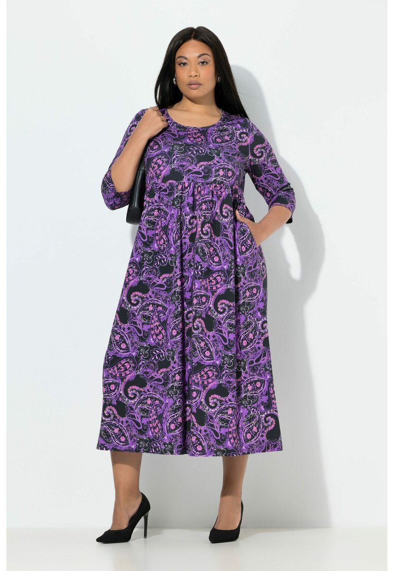 Violetti paisley-kuosinen mekko, jossa on kolme neljänneksen hihaa, laskeutuva siluetti ja taskut. Päällä mustat korkokengät.