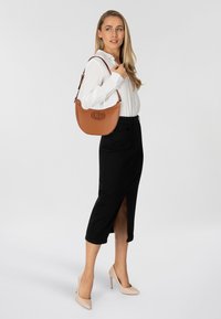 Borsa a mano in pelle marrone con forma curvata e logo prominente. Abbinata a una blusa bianca, una gonna nera e tacchi nudi.