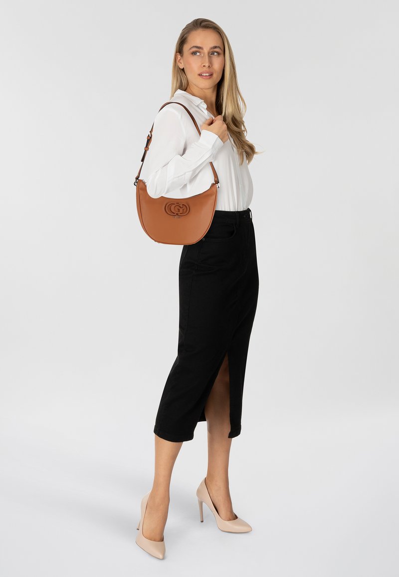 Borsa a mano in pelle marrone con forma curvata e logo prominente. Abbinata a una blusa bianca, una gonna nera e tacchi nudi.