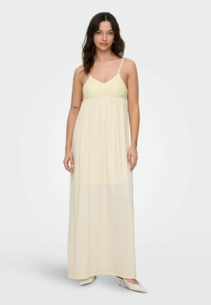 Kobieta w kremowej, bezrękawnikowej, długiej sukni maxi z górą wykończoną szydełkowaniem i cienkimi ramiączkami, stojąca na jasnym, jednolitym tle.