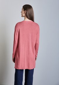 Top a maniche lunghe, svasato, in tessuto a maglia rosa chiaro, con orlo curvo e polsini a coste, indossato sopra pantaloni larghi blu scuro.