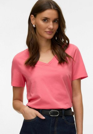 Woman with long brown hair wears a pink V-neck T-shirt, dark jeans with a black belt, and pearl stud earrings, standing against a white background.