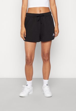 Kobieta ubrana w czarne szorty Nike, biały crop top, białe skarpetki Nike i białe sneakersy, stoi na tle jednolitego tła.