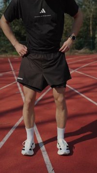 Δρομέας με μαύρο μπλουζάκι και σορτς Adidas, φορώντας λευκά αθλητικά παπούτσια και ρολόι, όρθιος σε έναν κόκκινο υπαίθριο στίβο.