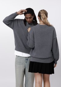 Un homme et une femme portant des pulls gris duveteux surdimensionnés ; l'homme fait face à l'avant avec la main sur la tête, la femme montre son dos avec une queue de cheval et une jupe noire.