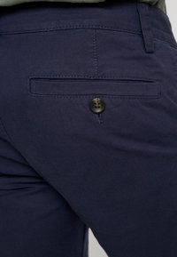 Pantaloni blu navy con una texture liscia, dotati di una tasca posteriore e una chiusura con bottone nero. Design semplice con dettagli di taglio su misura.