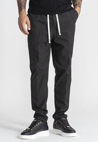 Hombre con pantalones negros ajustados de corte estrecho con cordones blancos y zapatillas negras de caña baja con suelas blancas, de pie con las manos en los bolsillos.