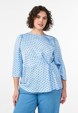 Žena s kučeravými vlasmi, oblečená v svetlomodrej blúzke s bodkami a 3/4 rukávmi a svetlomodrých nohaviciach na bielom pozadí.