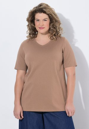 Femme aux cheveux bouclés portant un t-shirt marron à manches courtes et col en V, et un pantalon bleu foncé, debout devant un fond blanc uni.