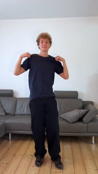 T-shirt preta de algodão com mangas curtas, ajuste solto, usada com calças pretas. Piso de madeira clara e sofá cinza ao fundo.