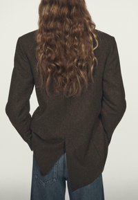 Osoba z długimi falującymi brązowymi włosami, ubrana w ciemno-brązową asymetryczną marynarkę i niebieskie dżinsy, stojąca z rękami w kieszeniach, tyłem do aparatu.