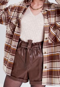 Femme portant une chemise à carreaux marron boutonnée, un haut en maille crème à col en V, un short en cuir marron avec ceinture, et un collier en chaîne dorée.