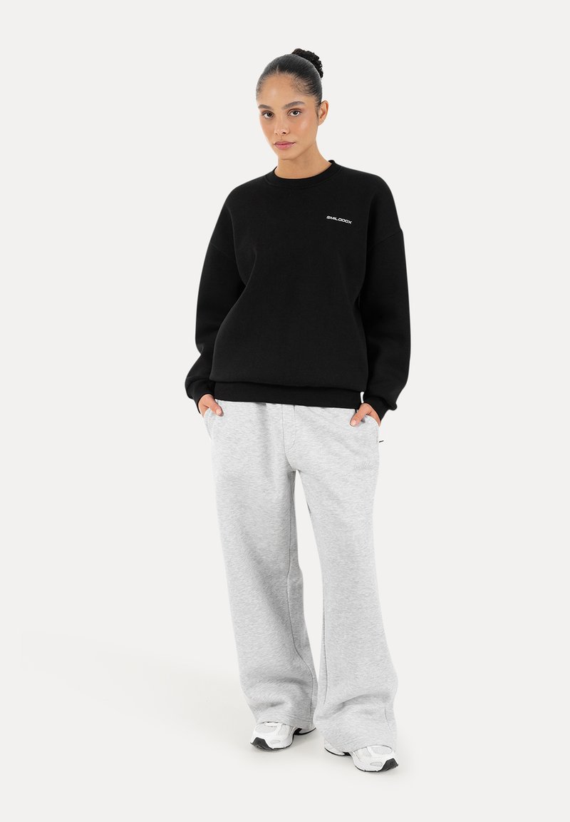 Femme debout, les mains dans les poches, vêtue d’un sweat-shirt noir, d’un pantalon large gris clair et de baskets blanches sur un fond uni.