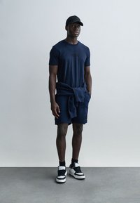 Marineblaues kurzärmeliges T-Shirt mit Logo, passende Shorts und eine schwarze Baseballkappe. Weiße und schwarze Turnschuhe vervollständigen das Outfit. Glatter Stoff, legere Passform.