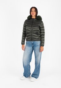 Giacca a vento verde con cappuccio foderato in pelliccia, motivo trapuntato, zip frontale, abbinata a jeans a gamba larga blu chiaro e sneakers bianche.