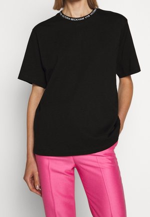 Femme portant un t-shirt noir avec un texte blanc sur le col et un pantalon rose vif, posant avec une main dans la poche.