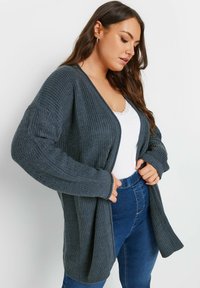 Cardigan lavorato a maglia di un grigio scuro con vestibilità ampia, apertura frontale e texture a costine. Maniche lunghe e tasche laterali, abbinato a un top bianco e jeans blu.