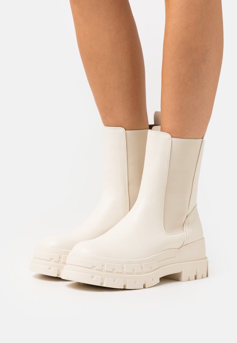 Bottes épaisses couleur crème mi-mollet avec semelles crantées épaisses portées sur des jambes nues, sur un fond clair uni.