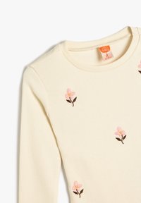 Helles, geripptes Langarmoberteil in Creme mit rosa stickten Blumen. Hat einen runden Halsausschnitt und eine weiche Textur. Pflegeetikett sichtbar.