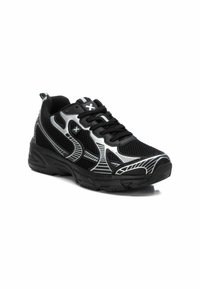 Zapatilla deportiva negra con acentos plateados, detalles en malla y suela acolchada. Presenta un diseño de cordones y superposiciones con patrones.