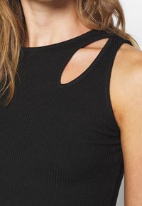 Gros plan d'une femme portant un haut noir sans manches côtelé avec une petite découpe près du col du côté droit.