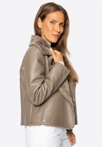 Beige leren jas met een pluizige grijze bontkraag, asymmetrisch ontwerp en met rits afgesloten zijzakken, met een zachte textuur.