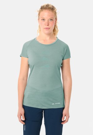 Donna con capelli ricci biondi, indossa una maglietta a maniche corte verde chiaro e pantaloni blu scuro, in piedi davanti a uno sfondo semplice.