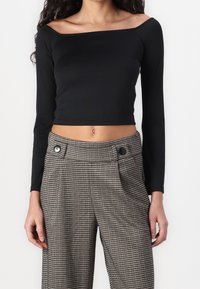 Schwarzes, kurz geschnittenes Langarmshirt mit schulterfreiem Ausschnitt, kombiniert mit hoch taillierten karomusternden Hosen mit kontrastierenden Knöpfen.