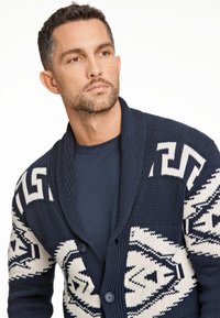 Mörkblå stickad kofta med sjalkrage, prydd med ett vitt geometriskt mönster, två knappar och en strukturerad yta.