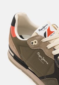 Olíva zöld sportcipő velúr és szövet anyagból, fekete és piros díszítéssel, krémszínű fűzővel és hangsúlyos Pepe Jeans logóval a nyelvén.