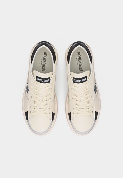 Ζευγάρι αθλητικά παπούτσια από δέρμα σε εκρού χρώμα με μαύρες λεπτομέρειες, κορδόνια και το λογότυπο "roberto cavalli" στις γλώσσες και τα εσωτερικά πέλματα, φαίνονται από πάνω.