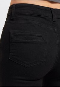 Svart denimjeans med en baklomme med subtile sømdetaljer og en glatt tekstur. Livremmen er synlig på toppen.