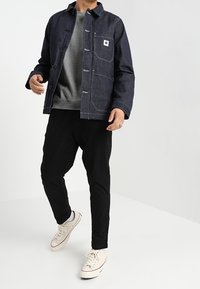 Tmavě modrá džínová bunda s zapínáním na knoflíky a kapsou na prsou, v kombinaci se šedou mikinou a černými zúženými kalhotami, doplněná bílými teniskami.