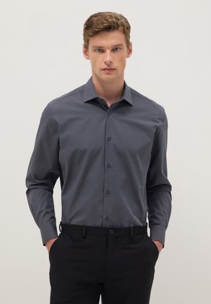 Homme portant une chemise habillée gris foncé boutonnée avec un pantalon noir, debout les mains dans les poches sur un fond clair uni.