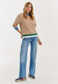 Beżowy sweter dzianinowy z niebieskimi i zielonymi akcentami, w zestawieniu z jasnoniebieskimi dżinsami w kształcie dzwonów oraz wielokolorowymi trampkami w wzory.